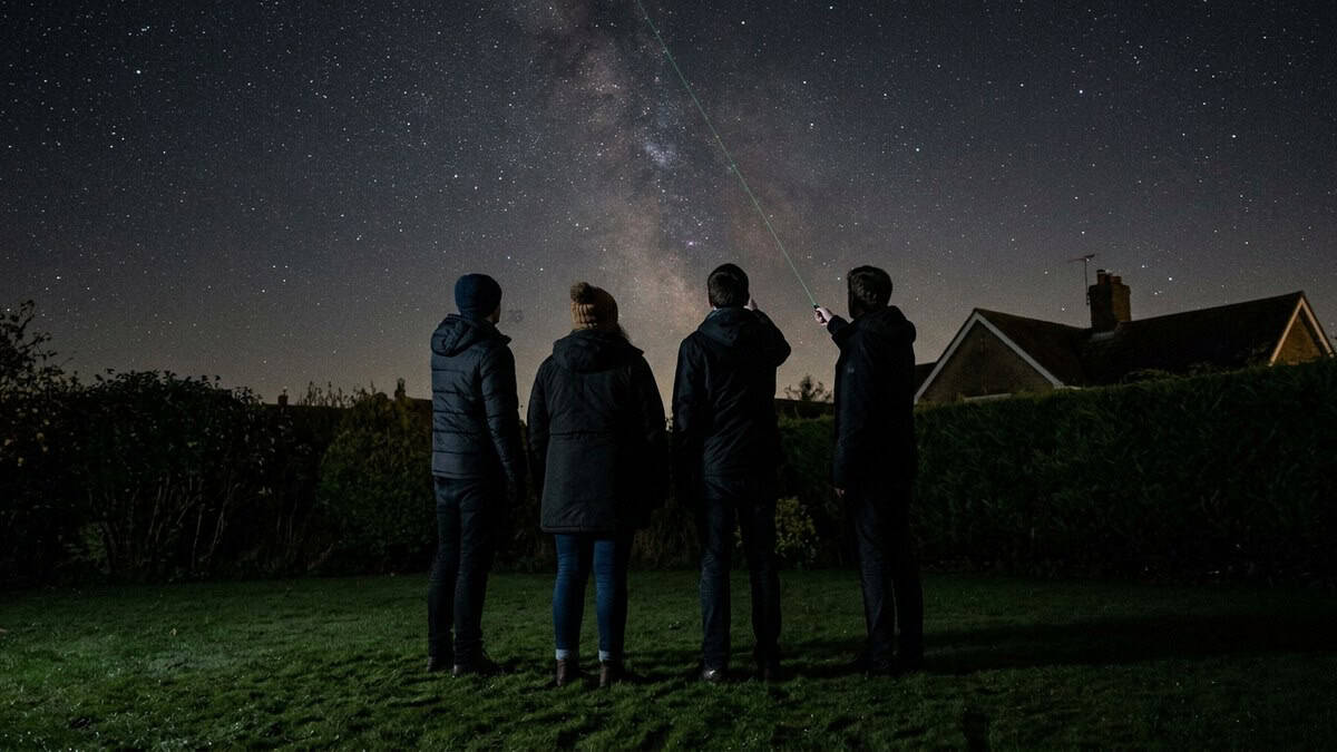 Small group standing on a dark lawn in October looking up as a thin green beam traces a constellation overhead